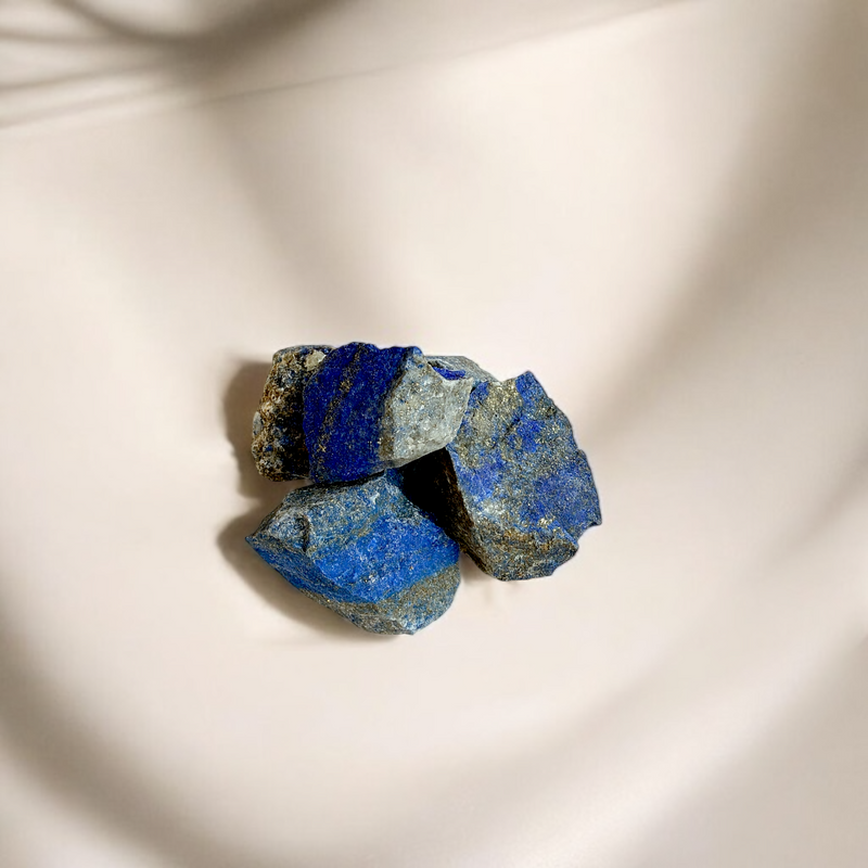 Raw Lapis Lazuli crystals in deep royal blue with gold and grey speckles, photographed on a soft beige background