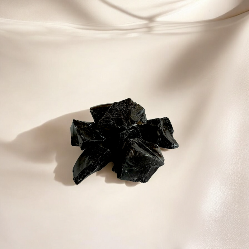 Raw Obsidian crystals in deep glossy black with sharp angular edges, photographed on a light beige satin-textured background