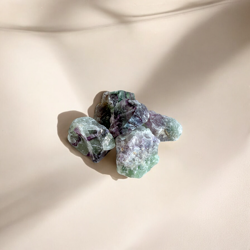 Raw Rainbow Fluorite crystals with shades of green, purple, and clear bands, photographed on a soft beige background