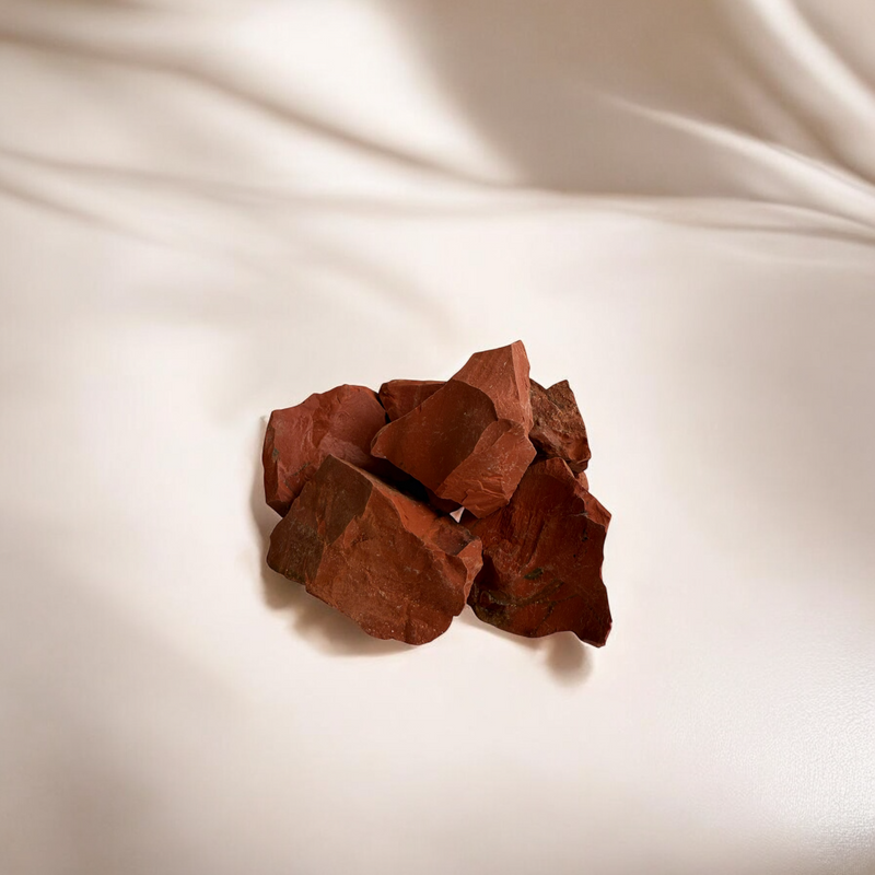 Cluster of raw Red Jasper stones in rich earthy tones with a smooth matte surface, placed on a creamy beige fabric background with soft shadows